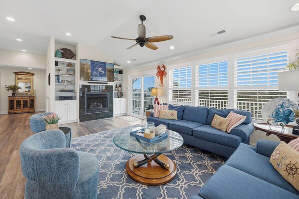 A spacious living room with a blue sofa, a glass coffee table, and a ceiling fan. - Koinania Cottage | 245 ft from the Beach | Private Pool (Virginia Beach)