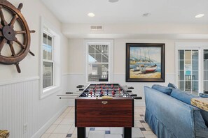 A room with a foosball table, a sofa, and a large ship's wheel on the wall. - Koinania Cottage | 245 ft from the Beach | Private Pool (Virginia Beach)
