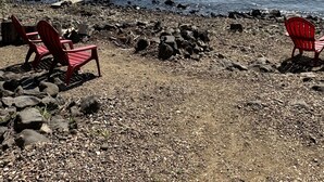 Una playa rocosa con dos sillas Adirondack rojas y un muelle de madera que se adentra en aguas tranquilas.