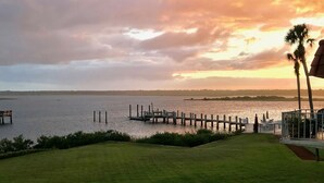A grassy area with a dock extending into a body of water, palm trees, and a distant shoreline. - Beach & Boating & Fishing Paradise! With 2 Kayaks Included!!! (St. Augustine)