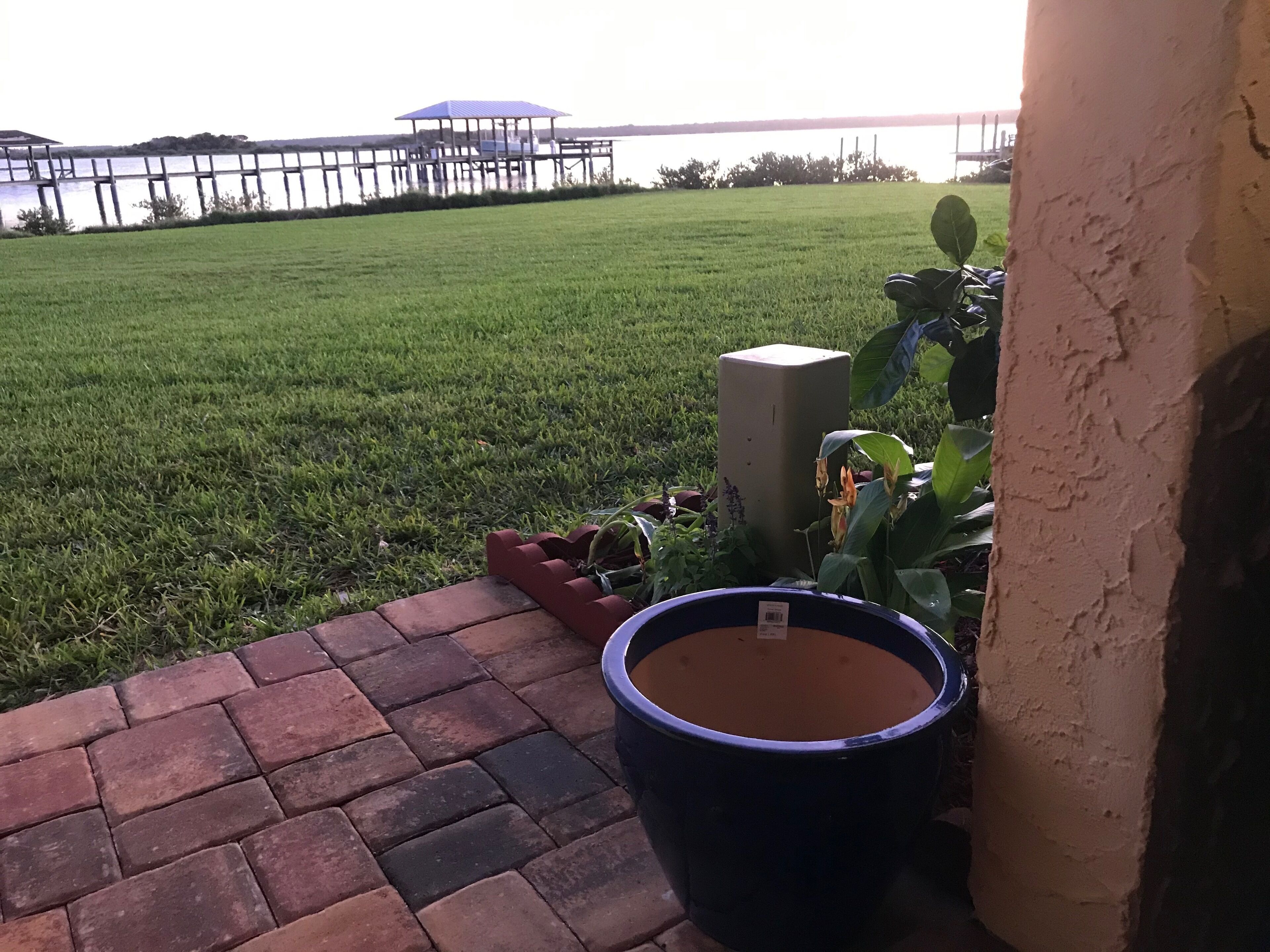 A grassy area with a dock in the background, a brick pathway, and a large planter.