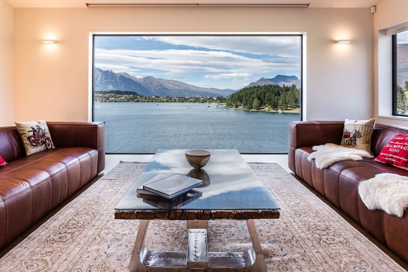 A living room with a large window offering a view of a lake and mountains. There are two leather sofas with decorative pillows, a coffee table with a bowl and books, and a patterned rug.