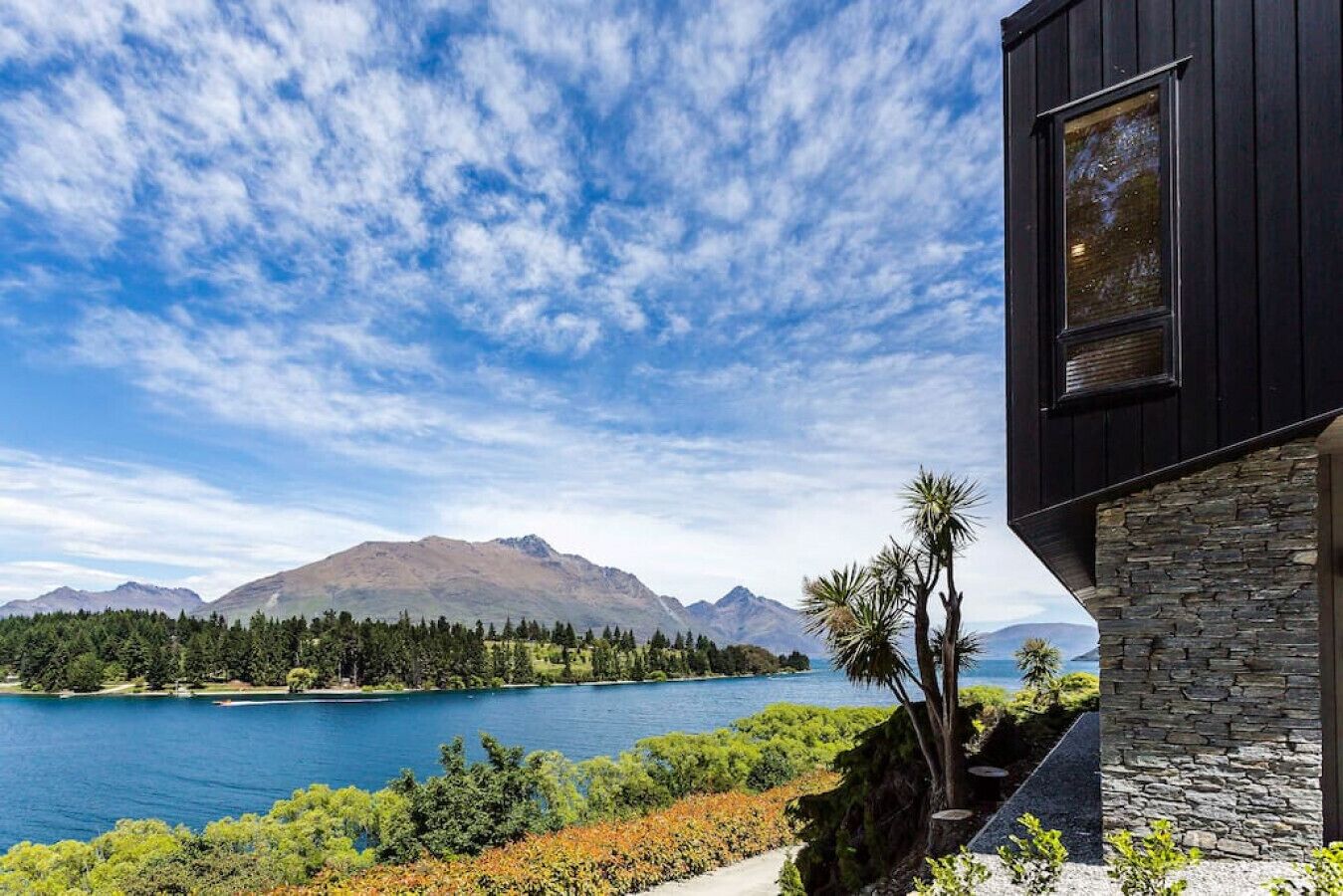A modern building with stone and wood exterior, overlooking a lake with mountains in the background.