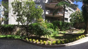 Un patio con un sendero de piedra, rodeado de árboles y arbustos, y un edificio con balcones al fondo.