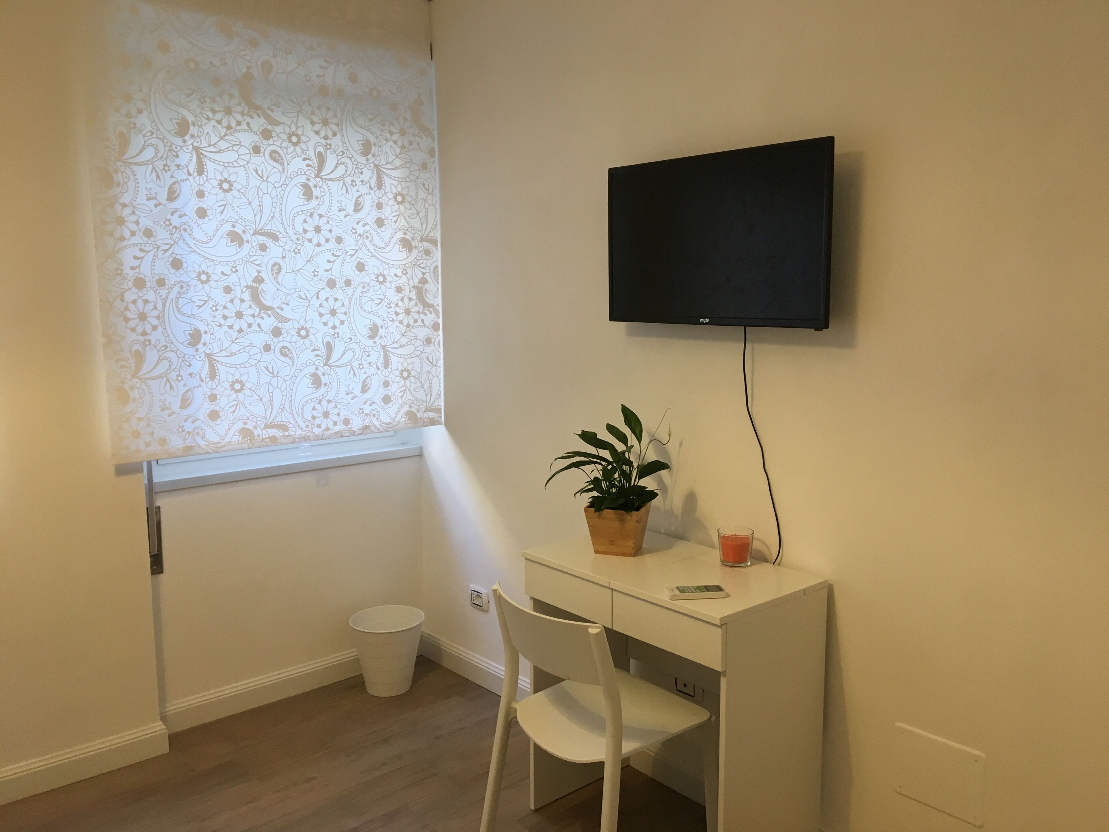 Et lille værelse med et hvidt skrivebord, en stol, en potteplante, et glas appelsinjuice, et fjernsyn på væggen, et vindue med blomstret gardin, og en hvid skraldespand.