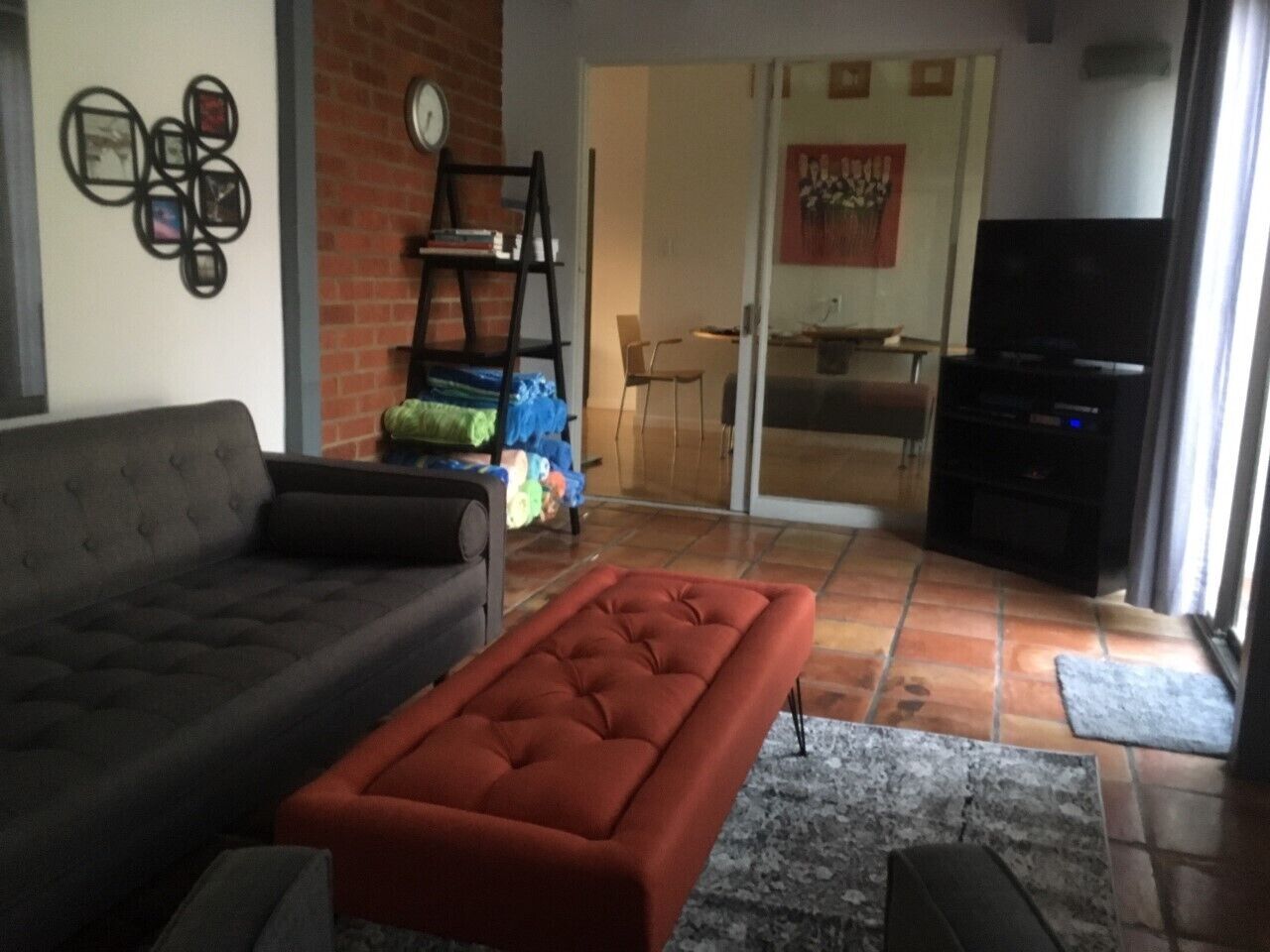 A living room with a grey sofa, a red ottoman, and a television. There is a brick wall and a door leading to another room.