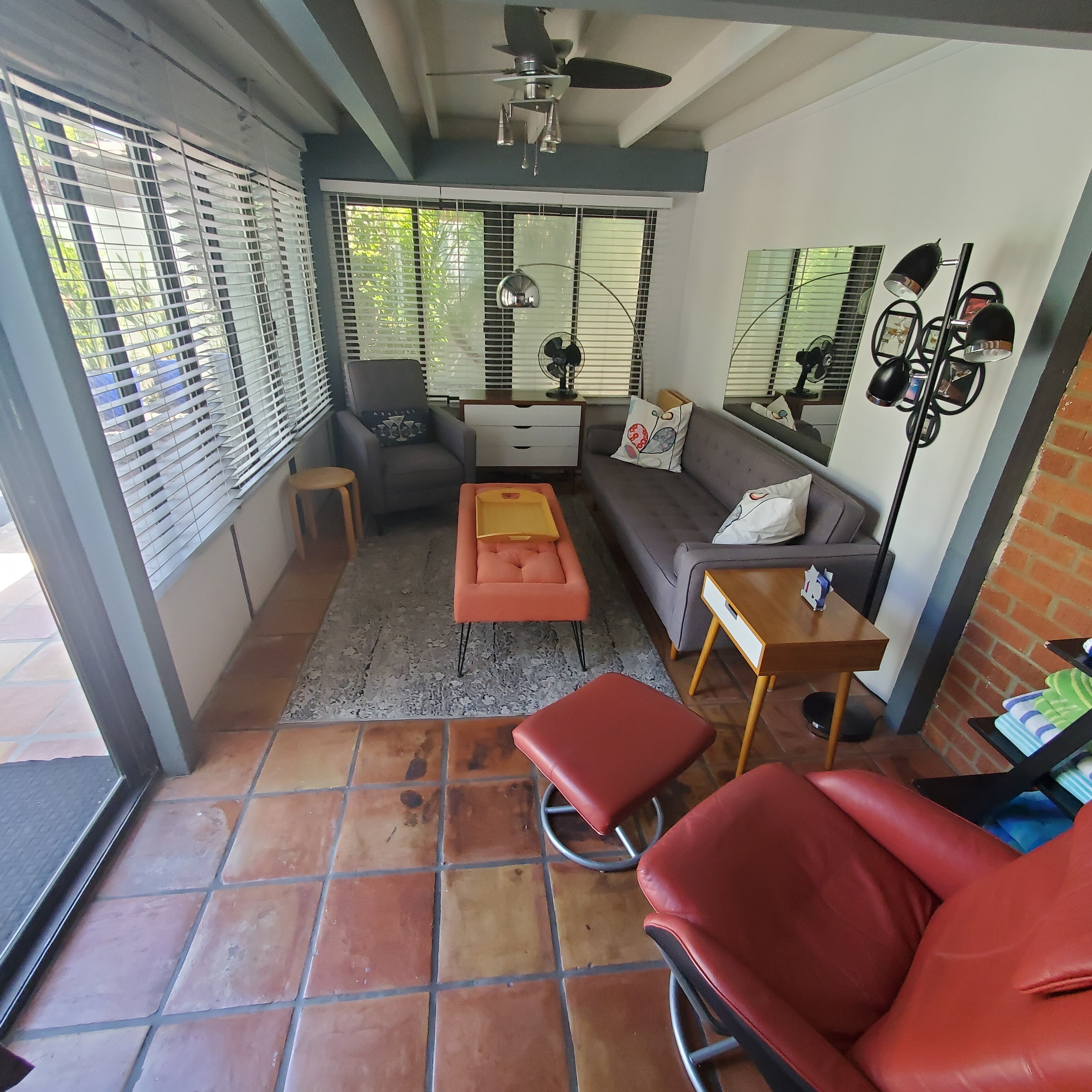 Ein Wohnzimmer mit einer Couch, einem roten Sessel, einem Couchtisch und einem kleinen Tisch mit einer Lampe. Es gibt Fenster mit Jalousien, einen Deckenventilator und eine Ziegelwand.