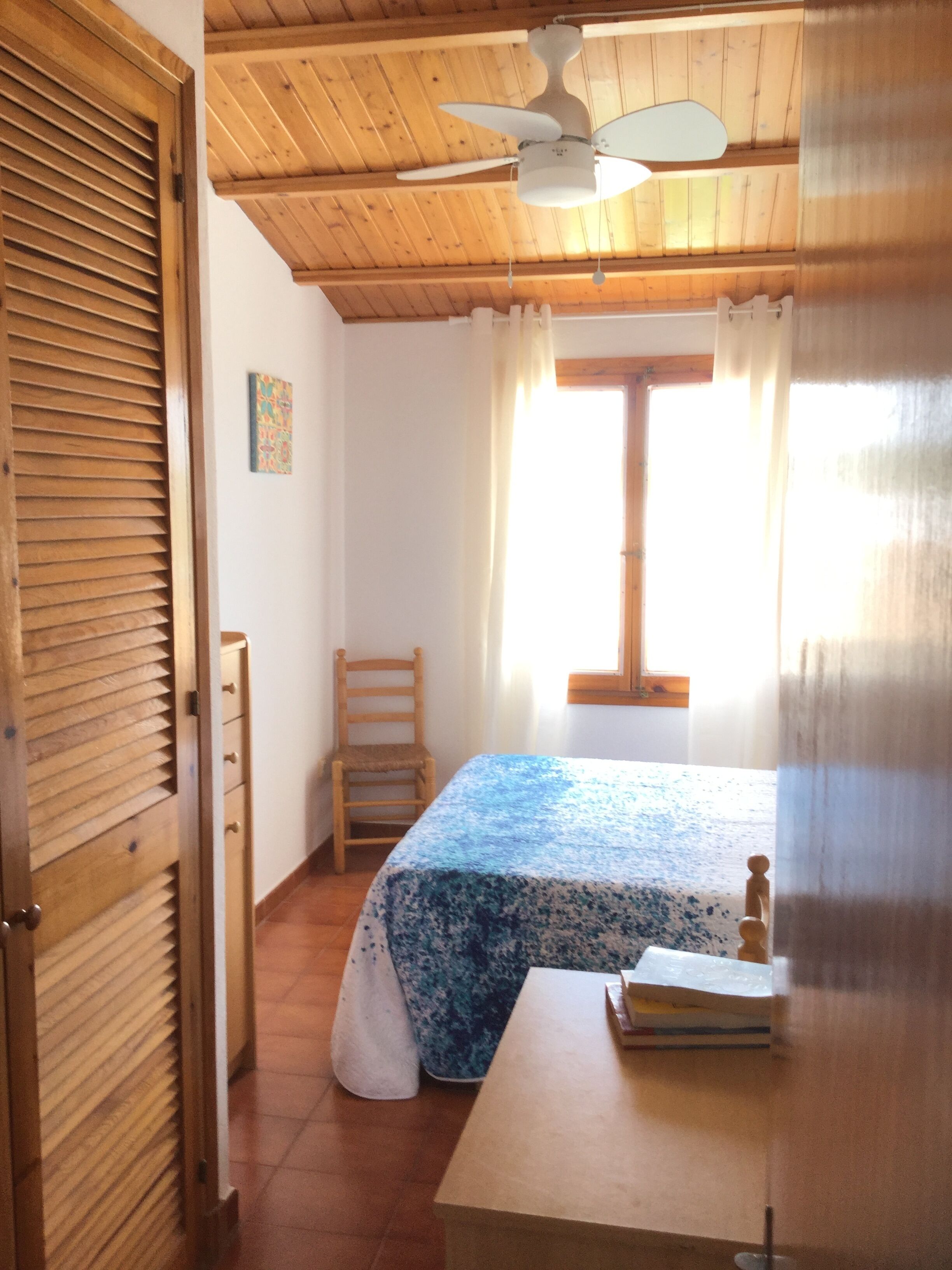 Une chambre à coucher avec un lit, une chaise, une table, un ventilateur de plafond et une fenêtre avec des rideaux.