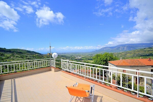 A terrace with a view of a hilly landscape, a white railing, and a single orange chair.