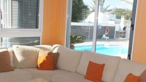 Un canapé d’angle beige agrémenté de coussins orange, une porte vitrée donnant sur un espace piscine et une petite table sur laquelle est posé un objet décoratif.