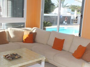 Un sofá seccional color beige con cojines naranja, una puerta de vidrio que da a una zona de piscina y una mesita con un objeto decorativo.