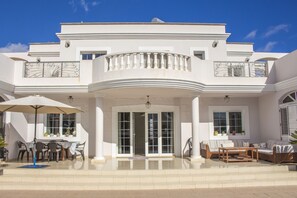 Una casa grande y moderna con un amplio patio amoblado, que incluye mobiliario exterior y una sombrilla grande.