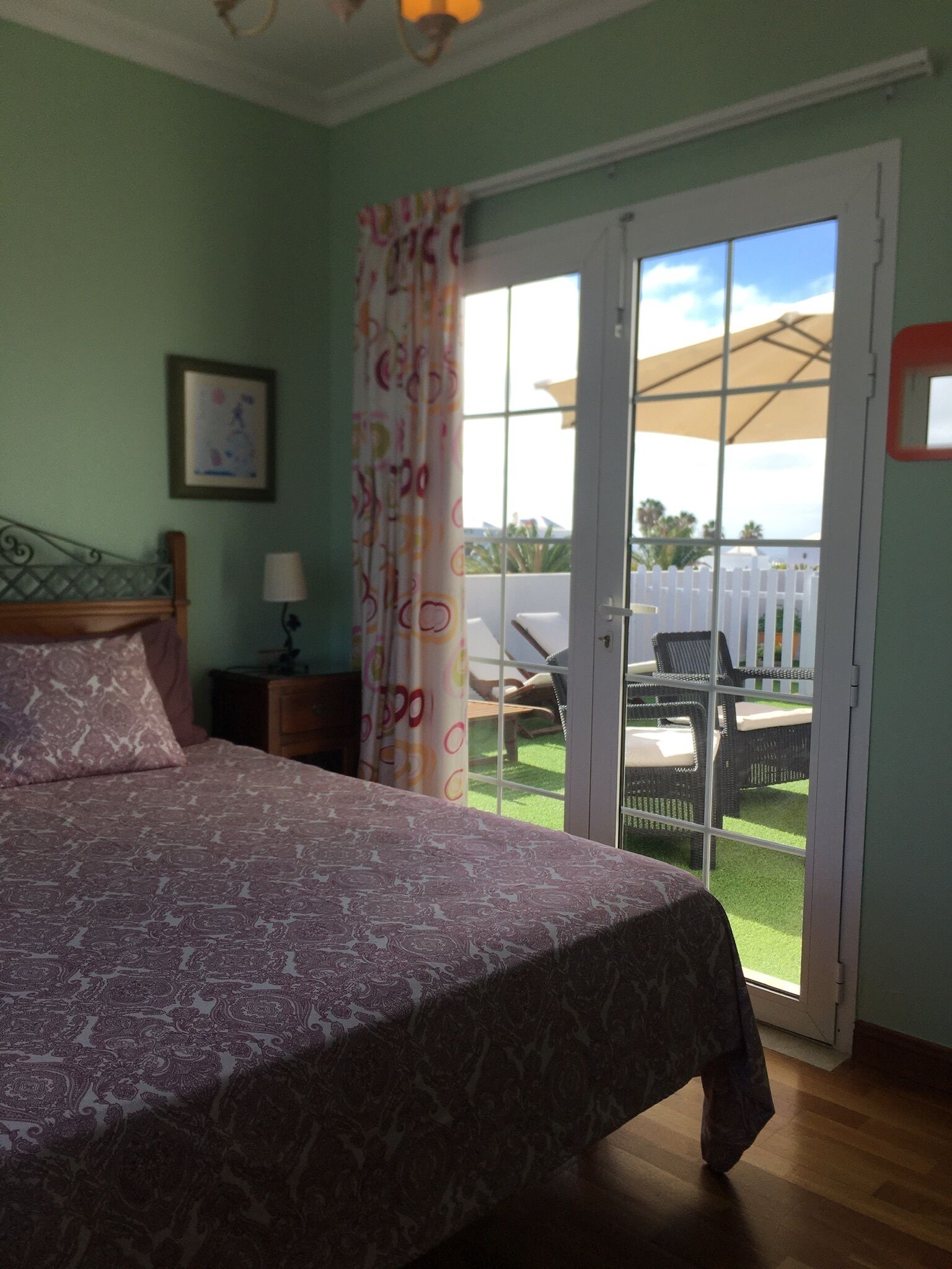 A bedroom with a bed, a nightstand, a lamp, a window with curtains, and a view of an outdoor patio with chairs and a table.