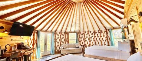 A yurt with a central skylight, a sofa, a bed, and a dining area with a TV and table.