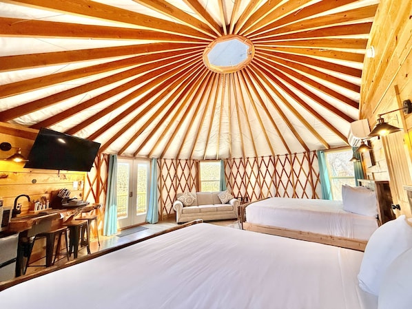 A yurt with a central skylight, a sofa, a bed, and a dining area with a TV and table.