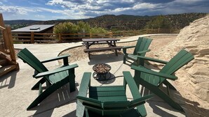 An outdoor seating area with green Adirondack chairs and a picnic table, surrounded by a wooden fence and a view of mountains and a building in the background. - Glamping Yurt near Zion with 2 King Beds & Views (Orderville)