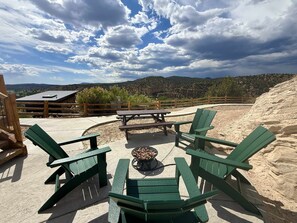 An outdoor seating area with green Adirondack chairs and a picnic table, surrounded by a wooden fence and a view of mountains and a building in the background. - Glamping Yurt near Zion with 2 King Beds & Views (Orderville)