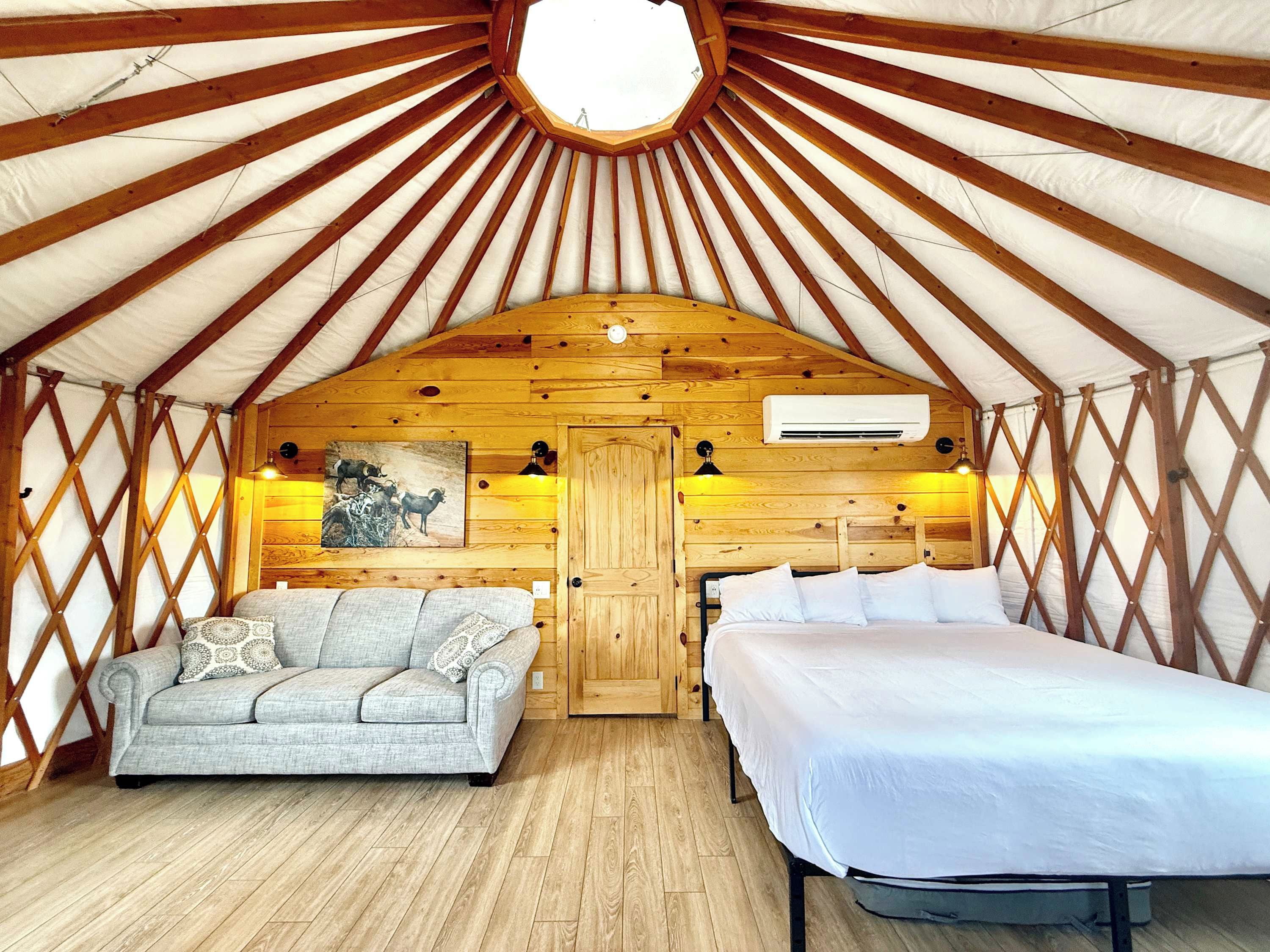 A cozy interior of a yurt with a wooden ceiling, a sofa, a bed, and a painting on the wall.