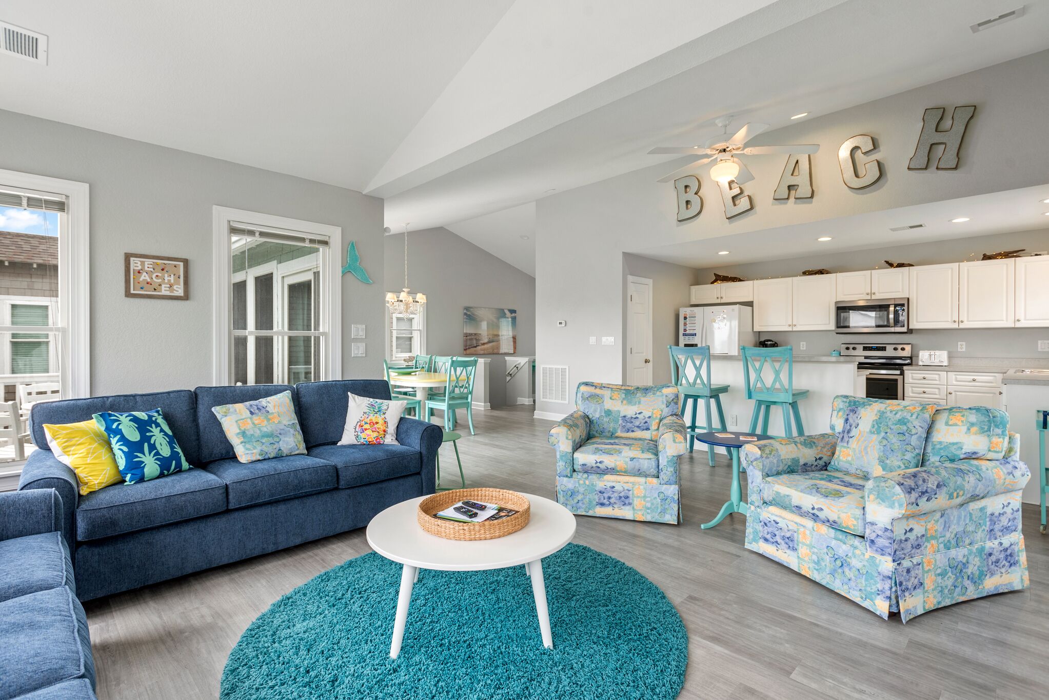 A spacious living room with a blue sofa, teal armchairs, and a white coffee table. A kitchen with white cabinets and a ceiling fan is visible in the background.
