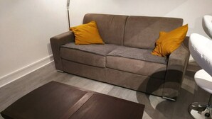 A modern living room with a sofa, two yellow cushions, and a glass coffee table. - Comfortable & affordable Parisian studio near the metro. (Ivry-sur-Seine)