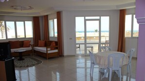 Uma sala de estar ampla com sofá, mesa de jantar e cadeiras, e grandes janelas com vista para a praia.