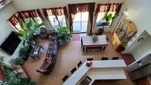 Amplia sala de estar con un sofá de cuero, una mesa de comedor y varias sillas. Hay varias plantas en macetas y un televisor montado en la pared.