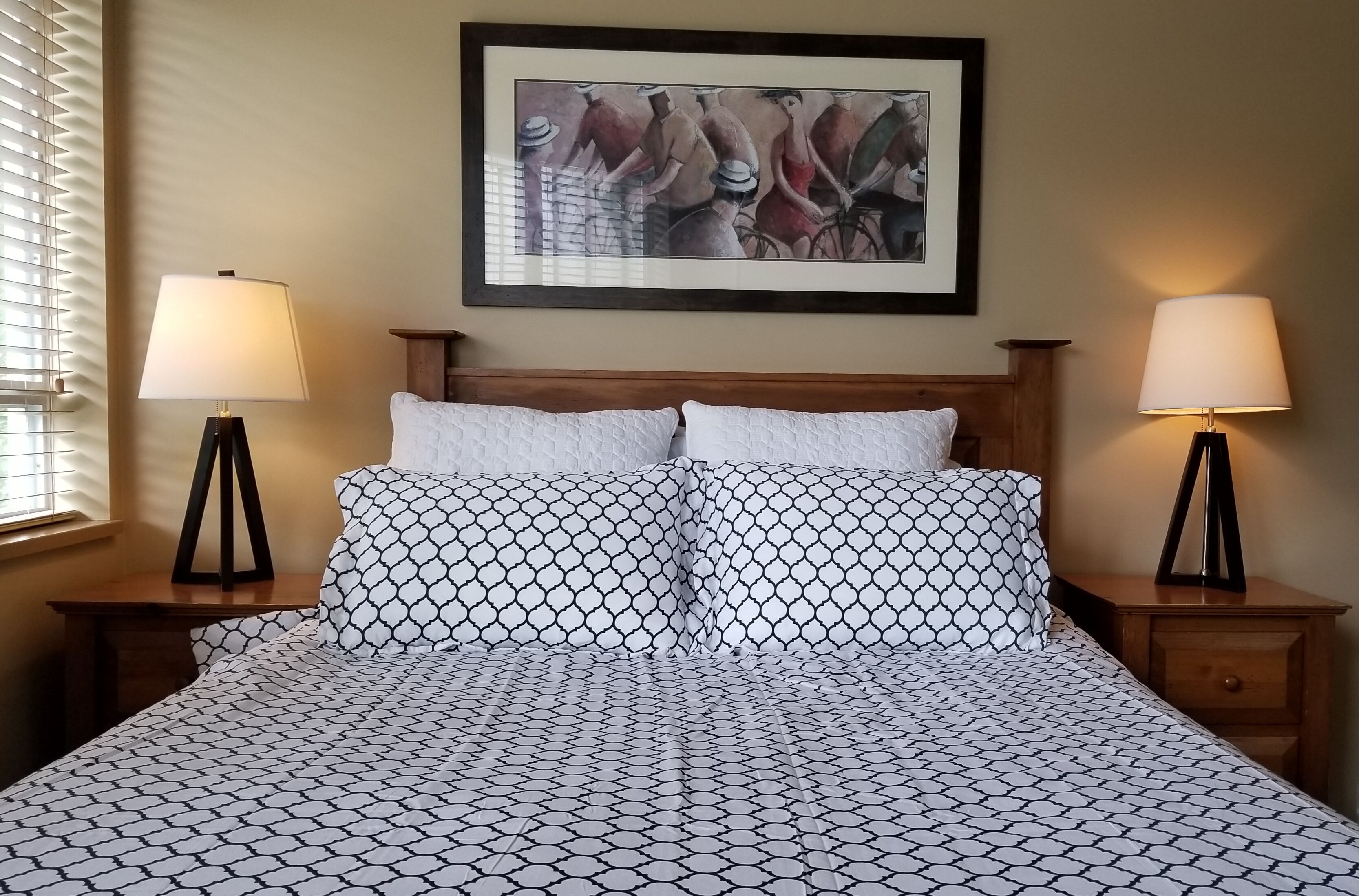 Een bed met een bedsprei met patroon, twee bedlampen, een houten hoofdbord, en een kunstwerk in een lijst erboven.