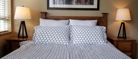 Een bed met een bedsprei met patroon, twee bedlampen, een houten hoofdbord, en een kunstwerk in een lijst erboven.