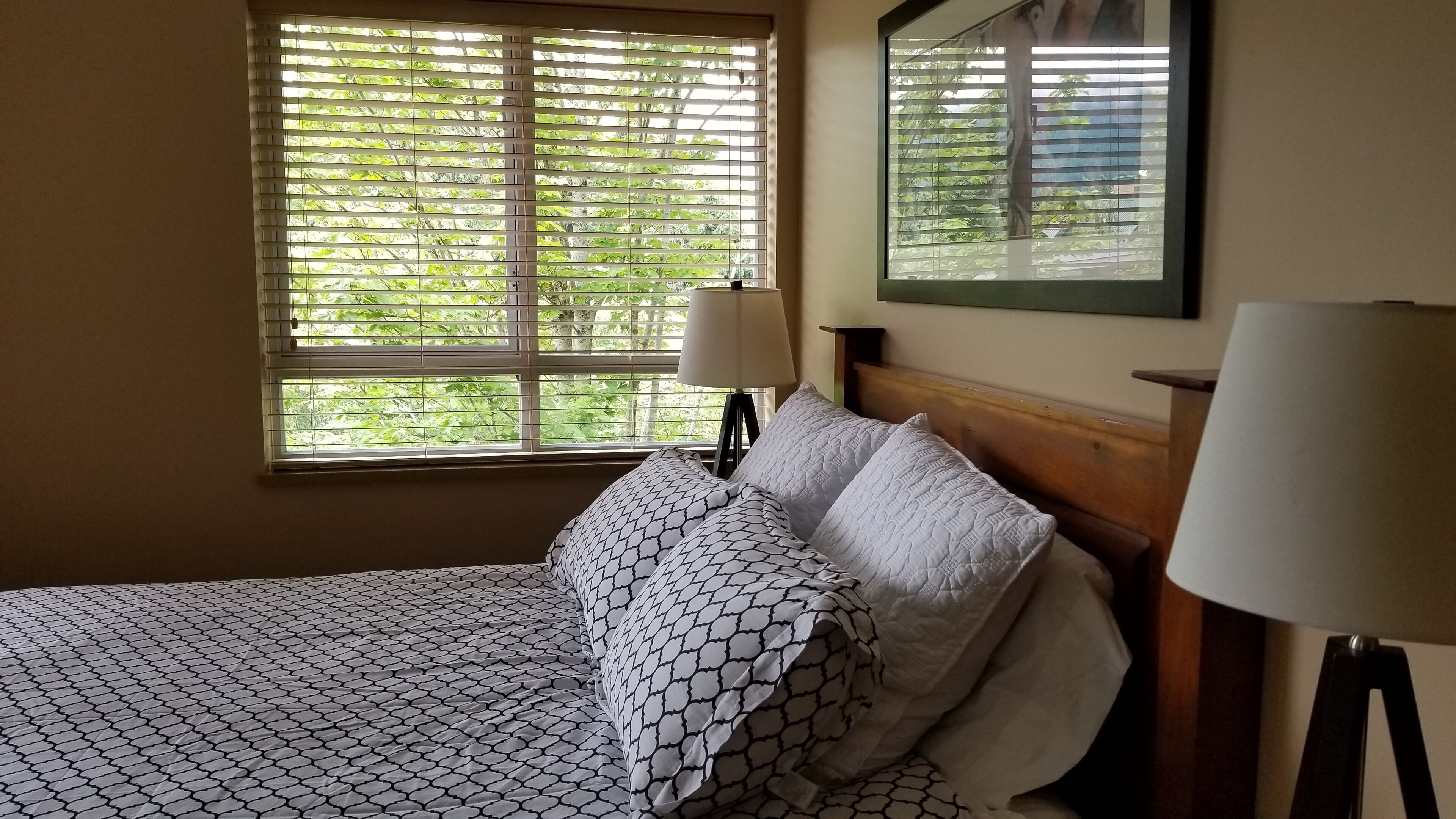 Ein Schlafzimmer mit Bett, gemusterten Betttextilien, einem Nachttisch mit Lampe, einem Fenster mit Jalousien und einem Spiegel an der Wand.