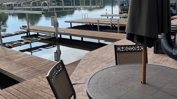 A wooden deck with a round table and chairs, overlooking a calm lake with a dock and a covered seating area.