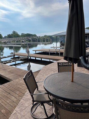 Una terraza de madera con una mesa redonda y sillas, con vista a un lago tranquilo que incluye un muelle y un área de estar cubierta.