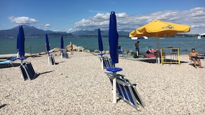 Ein Kiesstrand mit blauen und gelben Sonnenschirmen, Liegestühlen und freiem Blick auf das Meer und die dahinterliegenden Berge.