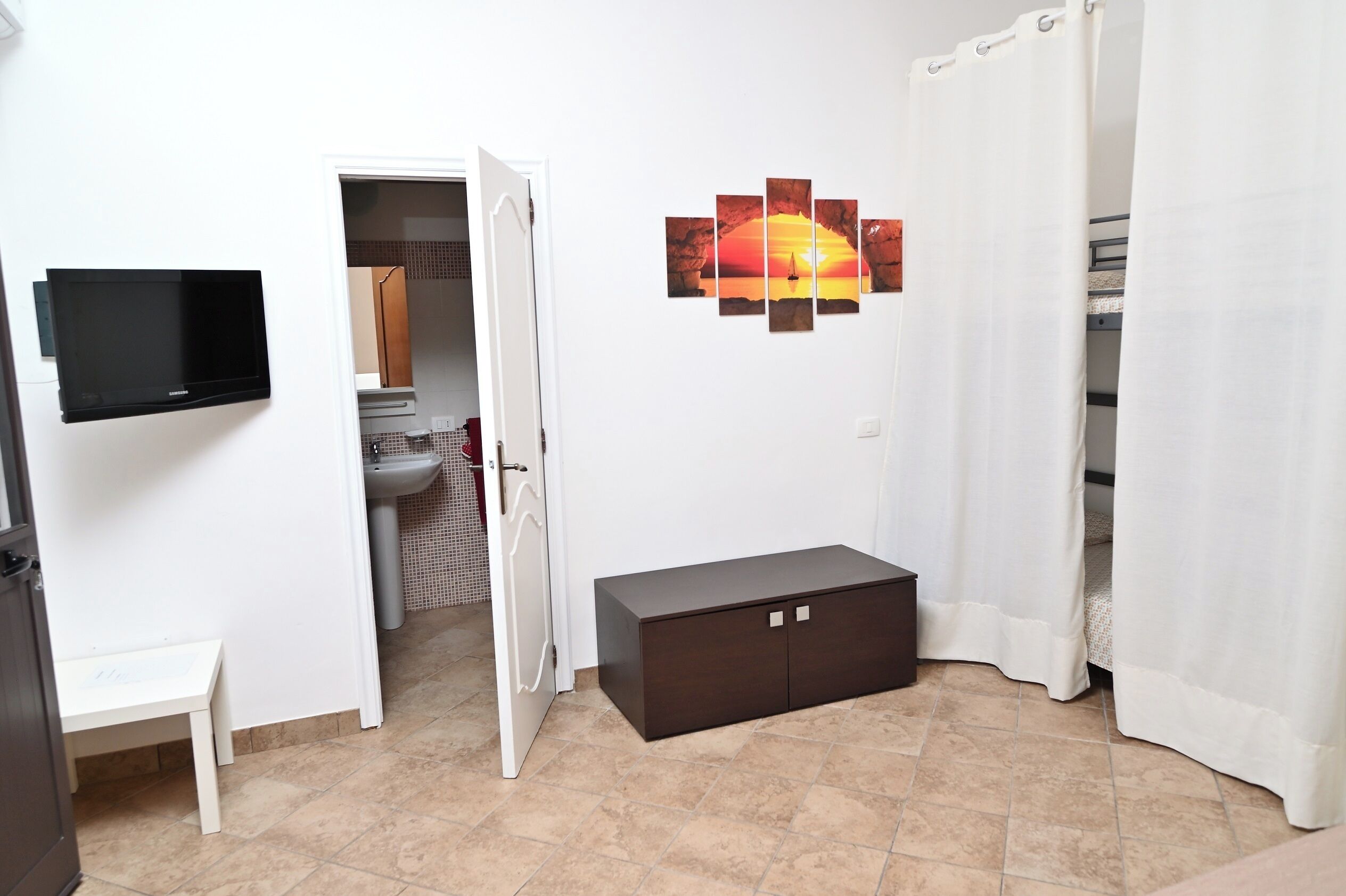 A room with a TV mounted on the wall, a cabinet, a bench, and a door leading to a bathroom.