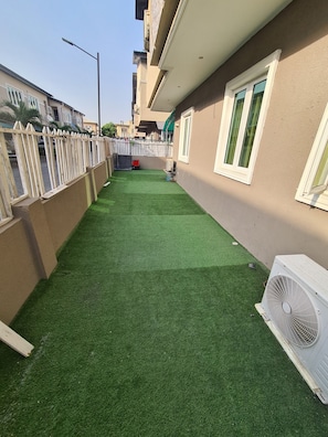 A narrow courtyard with artificial green turf, a white fence, and a building with a beige exterior and a window with green curtains. - Asa Apartments (Port Harcourt)