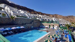 Un resort con piscina rodeada de edificios blancos, muros de piedra y sombrillas azules.