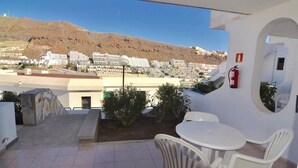 A balcony with a view of a mountainous landscape, white outdoor furniture, and a fire extinguisher on the wall.