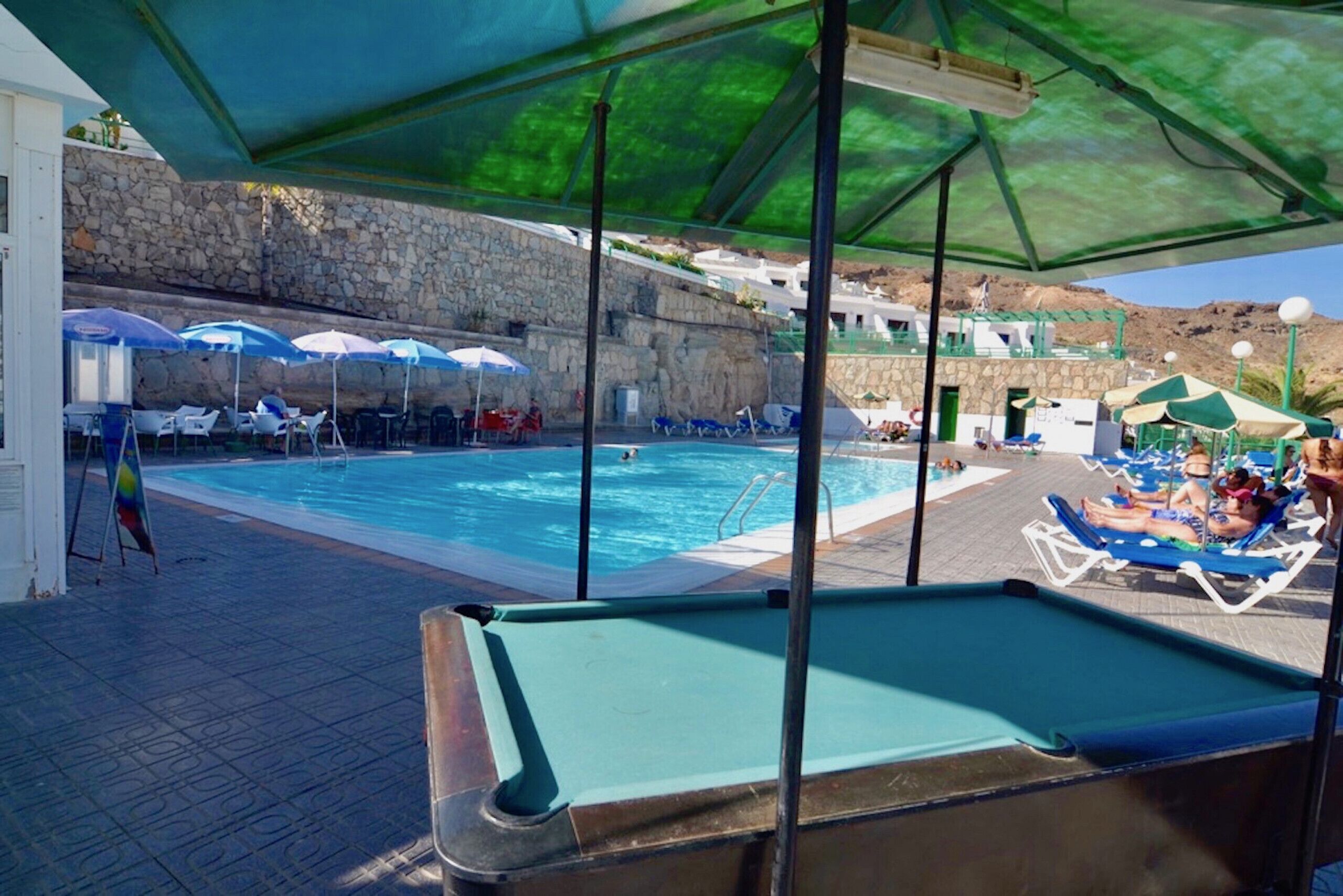 Un espace piscine avec des chaises longues et des parasols, une table de billard et un mur de pierre en arrière-plan.