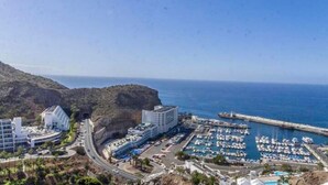Un puerto costero con varias embarcaciones amarradas, una carretera que lleva a un complejo de edificios y montañas al fondo.