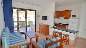 Una sala de estar con un sofá, una mesa de centro y un televisor. Hay una cocina con armarios de madera y un microondas.