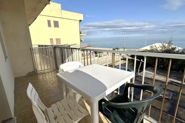 Ein Balkon mit Tisch und Stühlen, mit Blick auf eine Küstenlandschaft, im Hintergrund ein gelbes Gebäude.