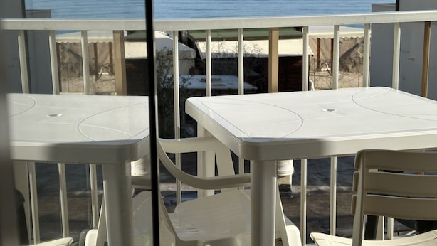 Uma varanda com cadeiras e mesas de plástico branco, com vista para a praia e o mar.