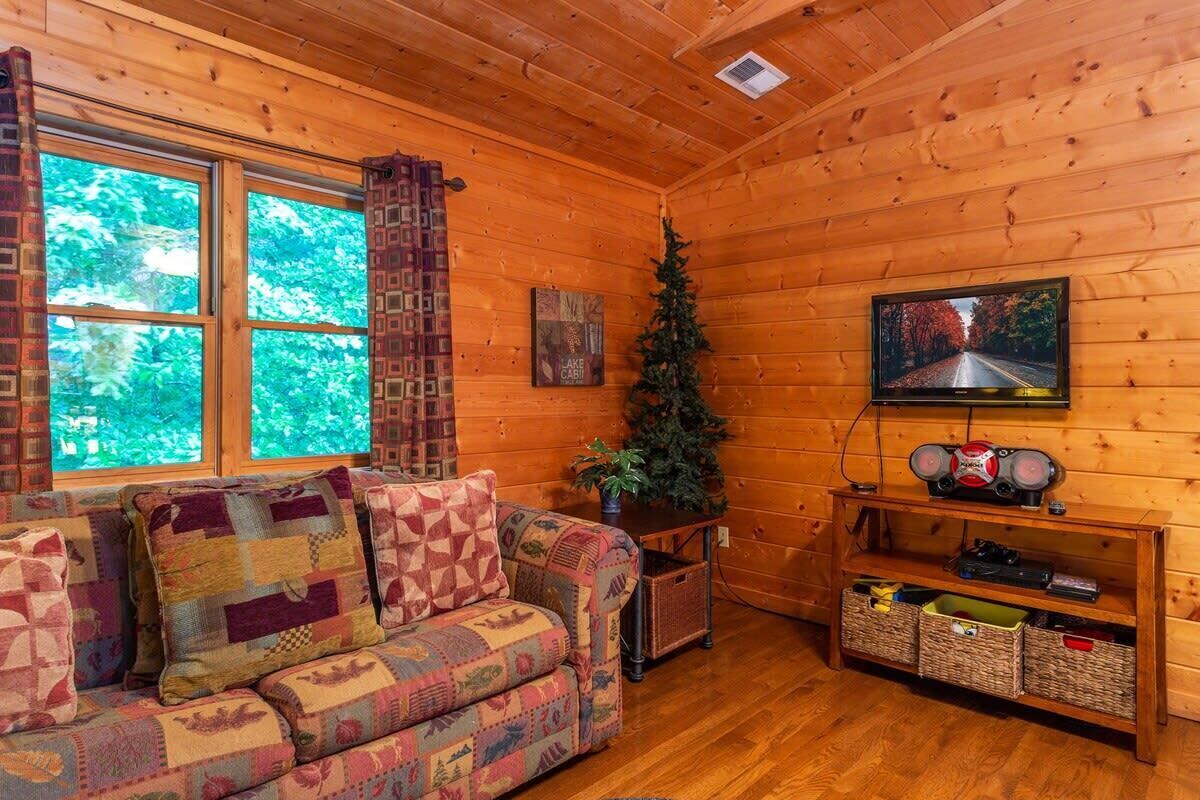A cozy cabin interior with a wooden floor, a quilted sofa, a television, and a Christmas tree.