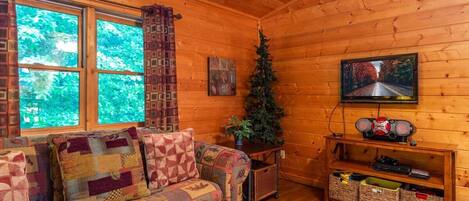 A cozy cabin interior with a wooden floor, a quilted sofa, a television, and a Christmas tree.