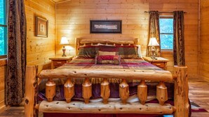 Camera da letto in una baita di legno con un letto al centro di una piattaforma, affiancato da comodini con lampade, quadri alle pareti e tende alle finestre.