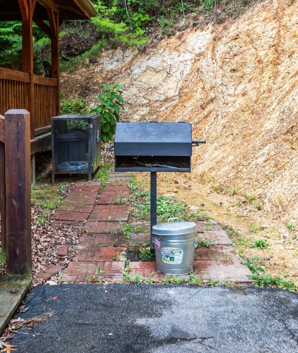 A barbecue grill and a trash can in a backyard with a wooden fence and a brick pathway.