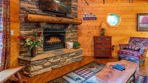 Un accogliente interno di una baita con camino in pietra, pareti in legno, TV a parete e una poltrona con motivo decorativo.