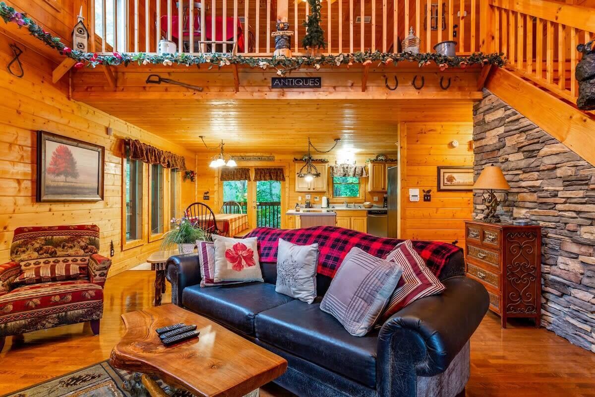 A cozy log cabin interior with a living room featuring a black leather sofa, a wooden coffee table, and a traditional armchair. A stone fireplace and a staircase with a wooden railing are visible in the background.
