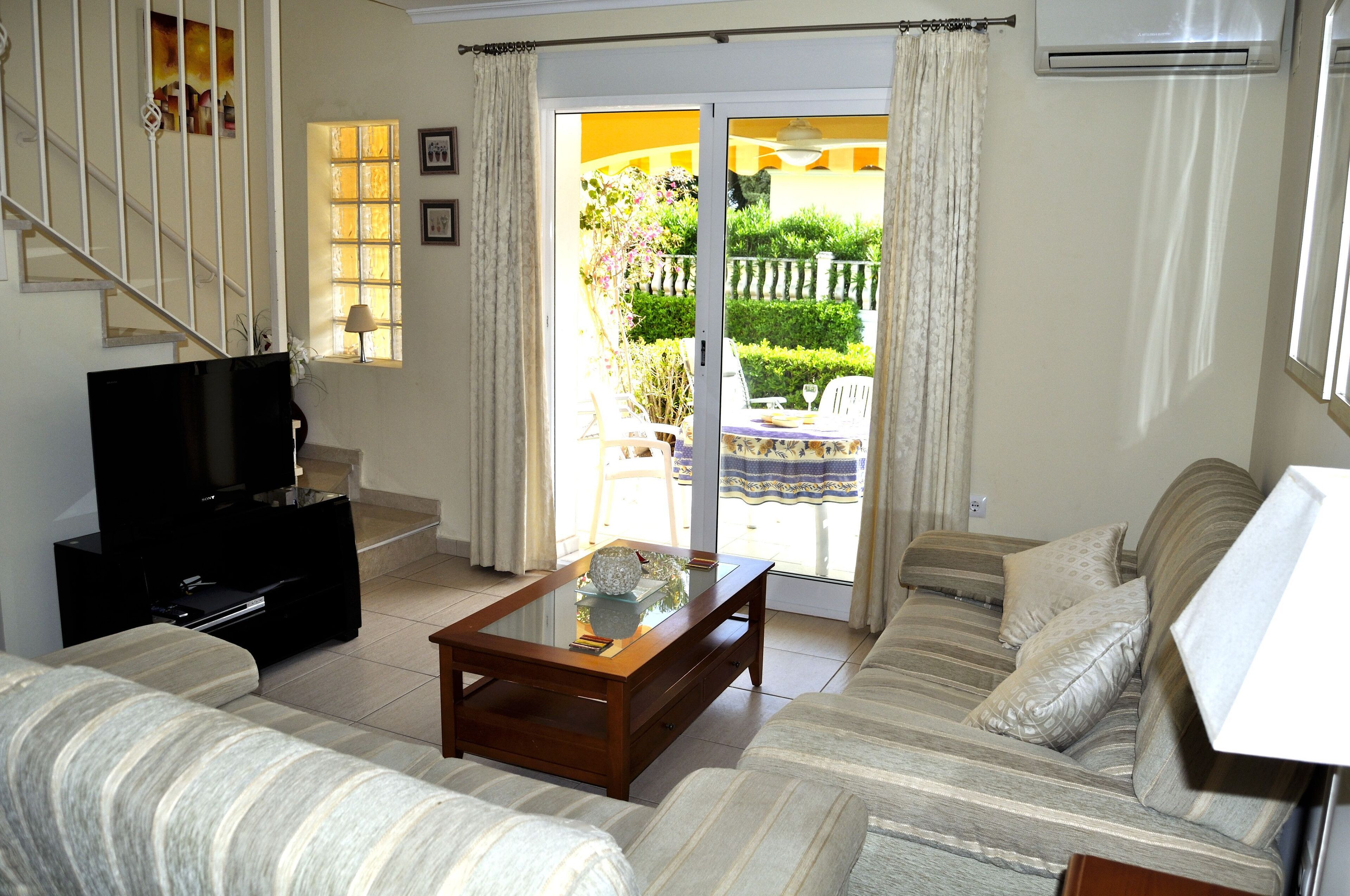 A living room with a sofa, a coffee table, a TV, and a sliding glass door leading to a garden.