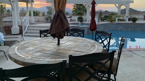Un espace aménagé au bord de la piscine, avec une table ronde et des chaises, une pergola recouverte d’un tissu drapé, et une vue sur les montagnes en arrière-plan.