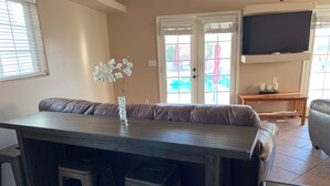 Un coin repas avec une table en bois, quatre tabourets, un vase avec des fleurs blanches, un téléviseur fixé au mur et une porte-fenêtre coulissante donnant sur l’extérieur.